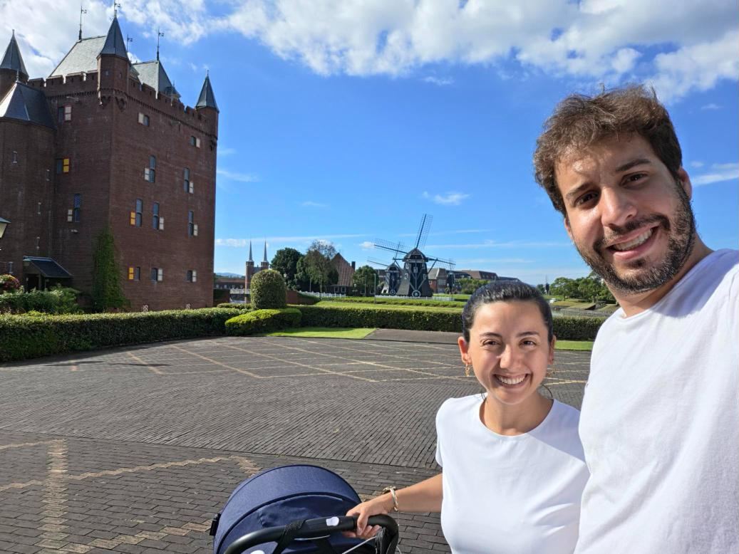 Rapha e Adam sorrindo com o carrinho do bebê em frente a um castelo e um moinho