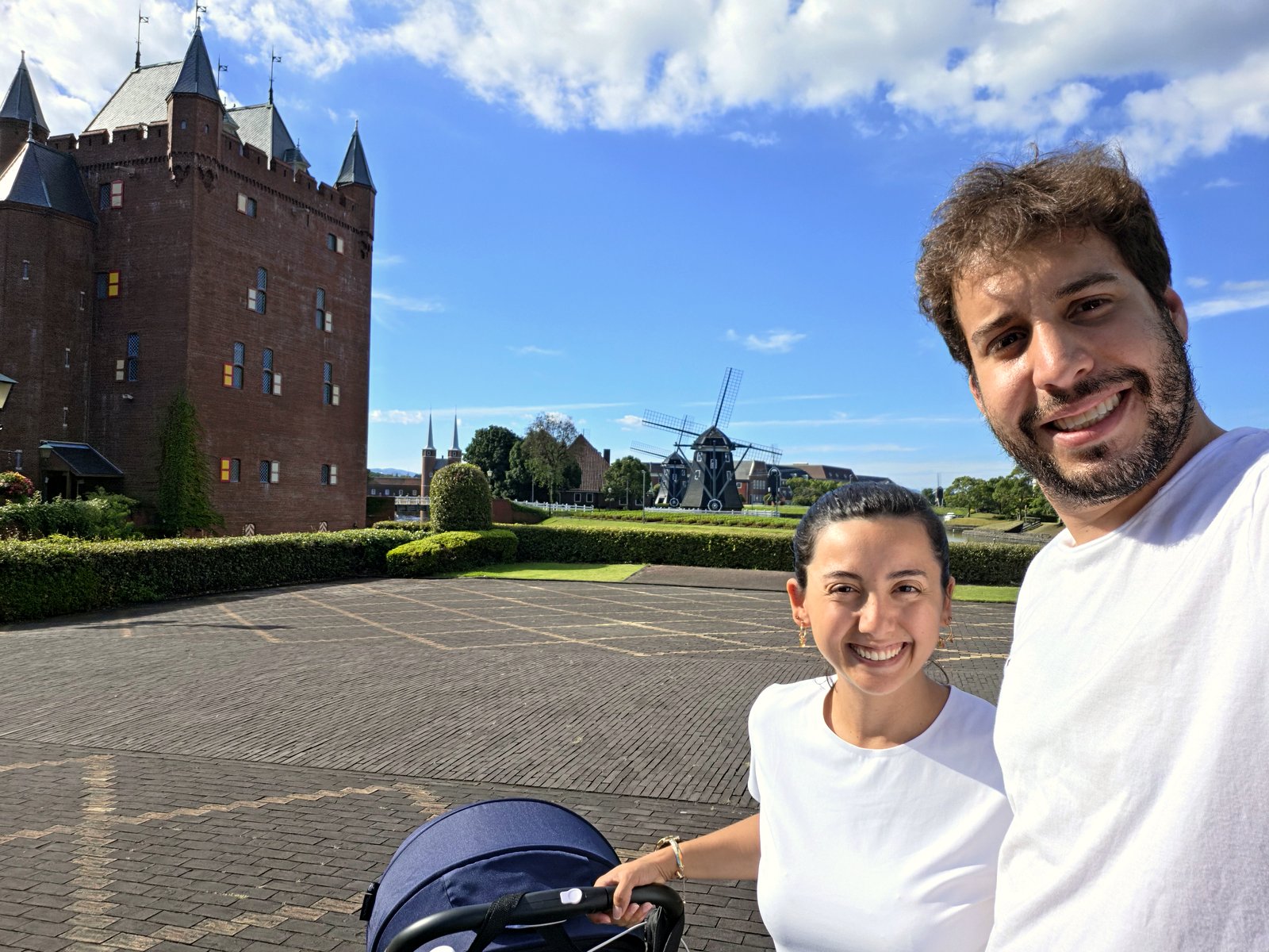 Rapha e Adam sorrindo com o carrinho do bebê em frente a um castelo e um moinho